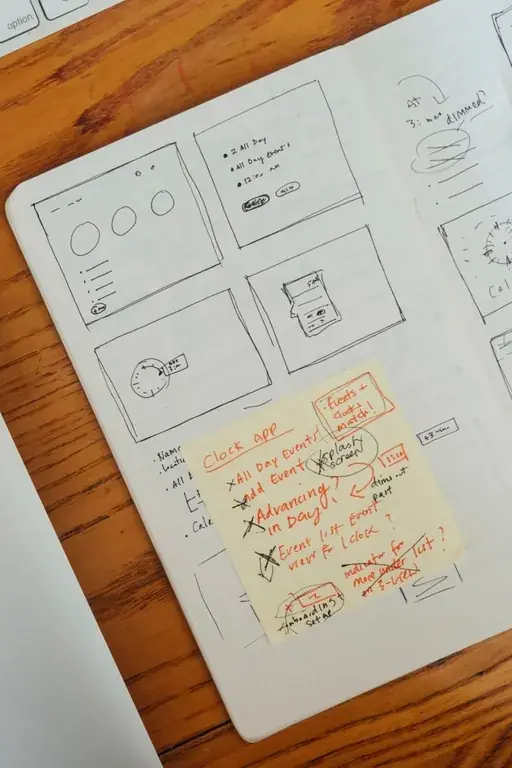 Close view of wireframe sketches and a sticky note covered in red handwritten notes and crossings-out